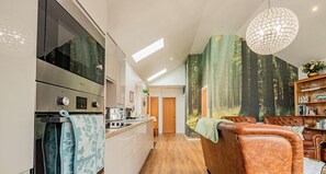 A modern kitchen with built-in appliances, a dining area with leather chairs, and a large window with a forest view. - Host & Stay | Little Highlands (Tenbury Wells)