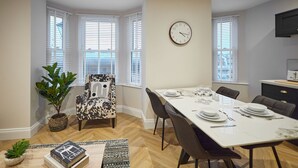 Un comedor con una mesa blanca dispuesta para cuatro, un sillón estampado y una planta en maceta.