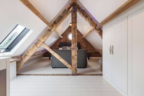 Ein Schlafzimmer im Dachgeschoss mit freiliegenden Holzbalken, einem grauen Sofa, einem runden Fenster und weißen Schränken.
