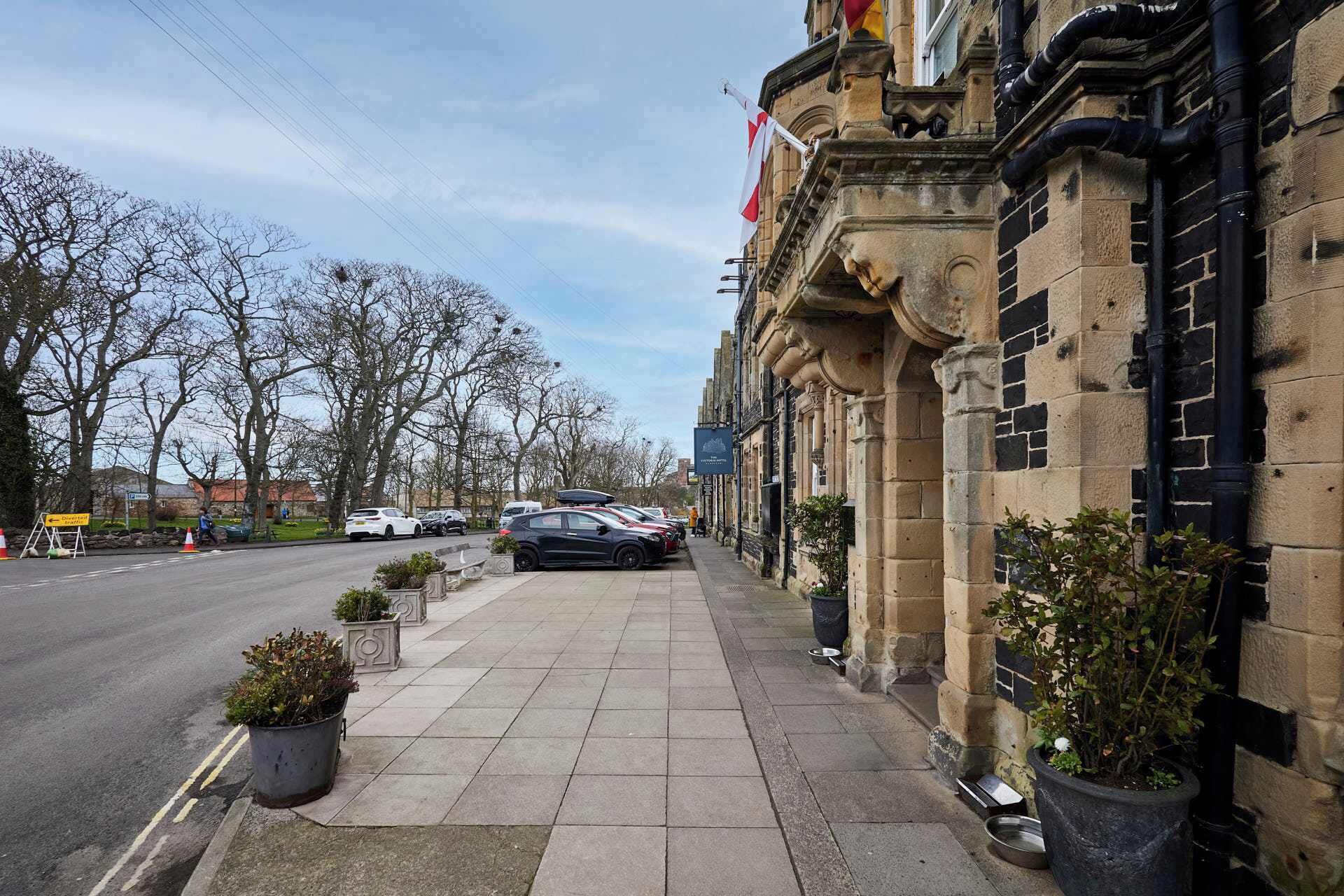 A street view with parked cars, a sidewalk, and a building with stone architecture.