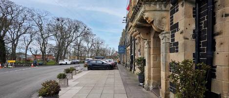 A street view with parked cars, a sidewalk, and a building with stone architecture.