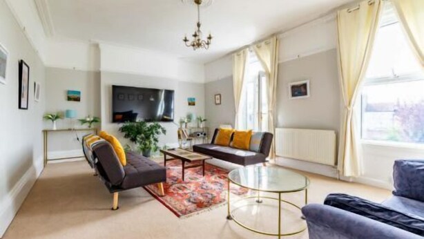 Ein modernes Wohnzimmer mit Sofa, Sessel und Couchtisch, einem Flachbildfernseher und einem großen Fenster mit Vorhängen.