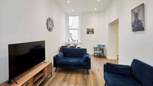 Ein Wohnzimmer mit Fernseher, zwei blauen Sofas, einem Holz-TV-Schrank, einer Uhr und einem Bild an der Wand.
