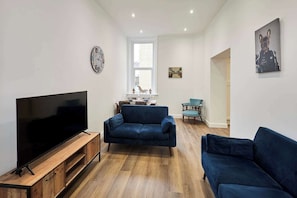Ein Wohnzimmer mit Fernseher, zwei blauen Sofas, einem Holz-TV-Schrank, einer Uhr und einem Bild an der Wand.