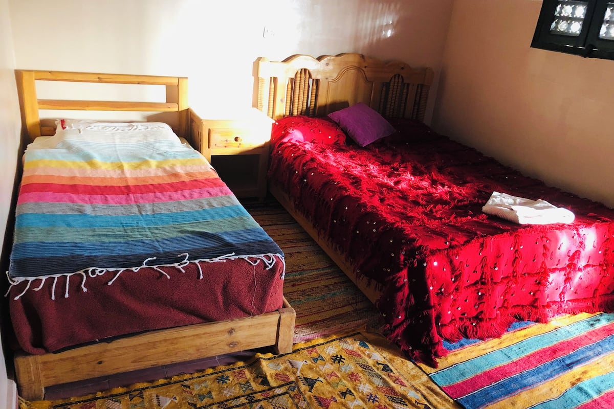 Ein kleiner Raum mit zwei Holzbett, eines mit einer gestreiften Decke und das andere mit einer roten Decke.