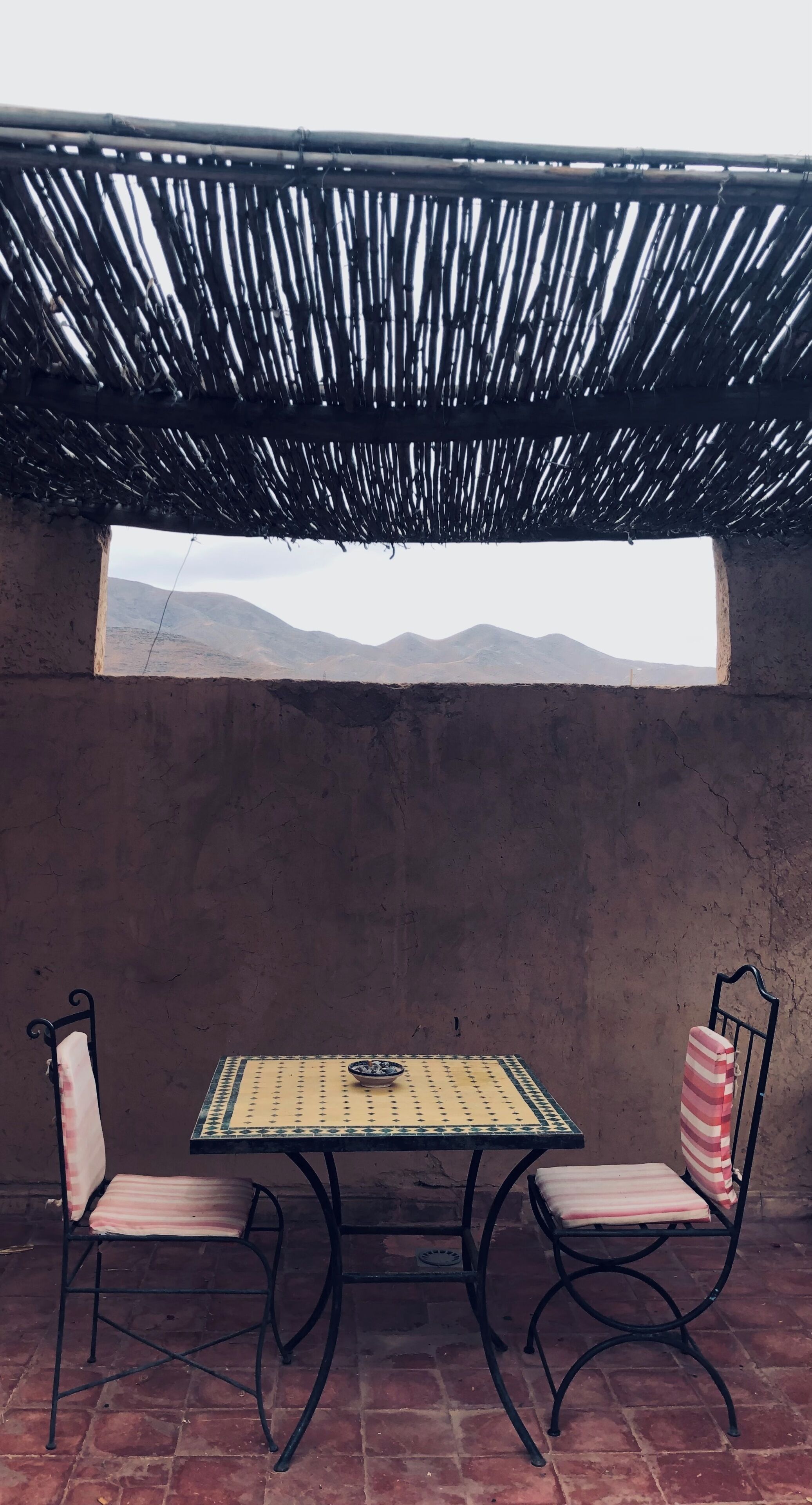 A rustic outdoor seating area with a table and chairs, a thatched roof, and a view of distant mountains through a window.