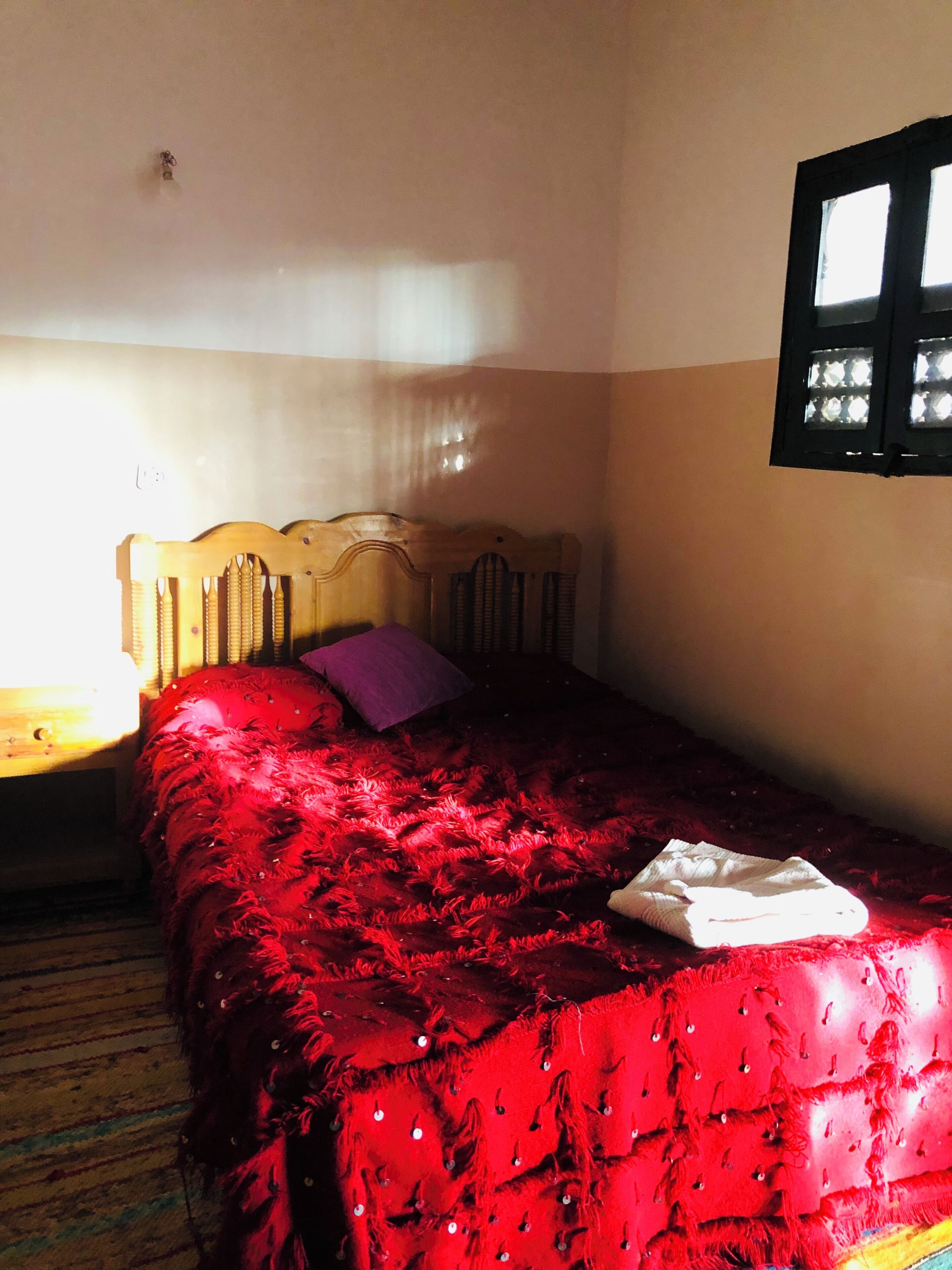 A single bed with a red bedspread, a wooden headboard, a small window, and a wall socket.