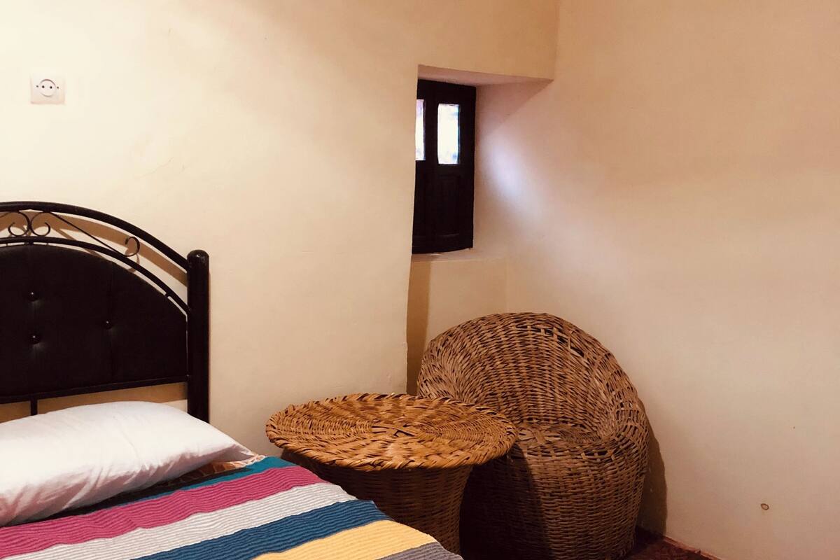 Ein Bett mit einer gestreiften Decke, ein Korbstuhl und ein kleines Fenster.