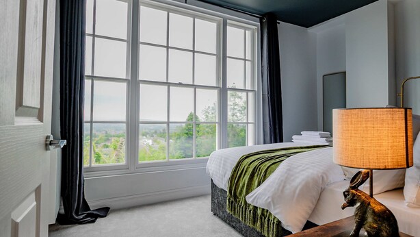 Ein Schlafzimmer mit großem Fenster, einem Bett mit weißer Bettwäsche, einem Nachttisch mit Lampe und einer Statue.