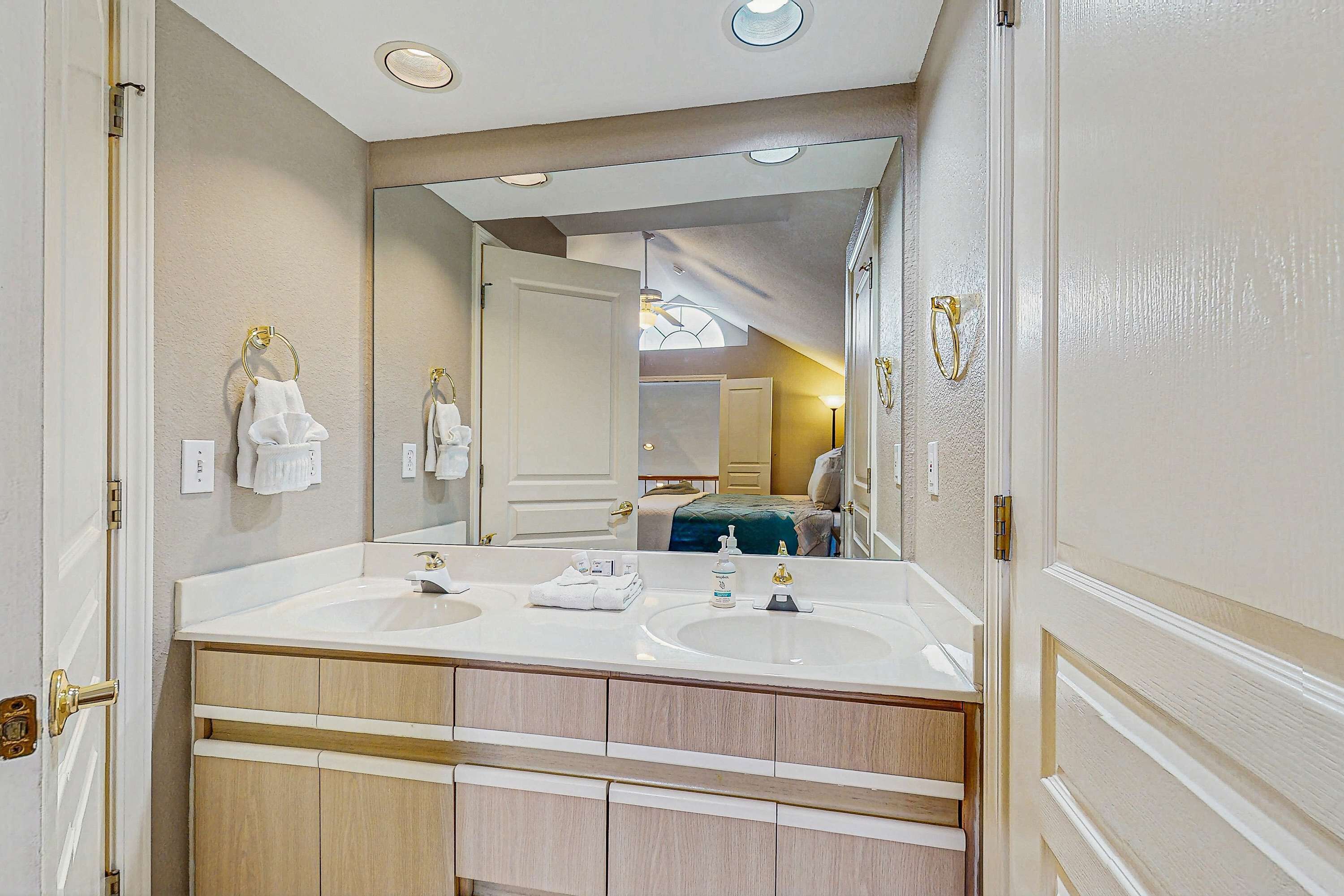 A bathroom with two sinks, a large mirror, and a view into the bedroom.