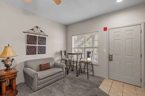 A living room with a grey sofa, a small table, two chairs, a decorative wall piece, and a door with a red light. - Spacious 2BR Condo - GOLF VIEWS - Pool & Dogs ok (Branson)