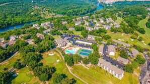 An aerial view of a residential area with houses, a swimming pool, tennis courts, and a dam in the background. - Pointe Royale - 1BR/1B - pools and fitness center (Branson)