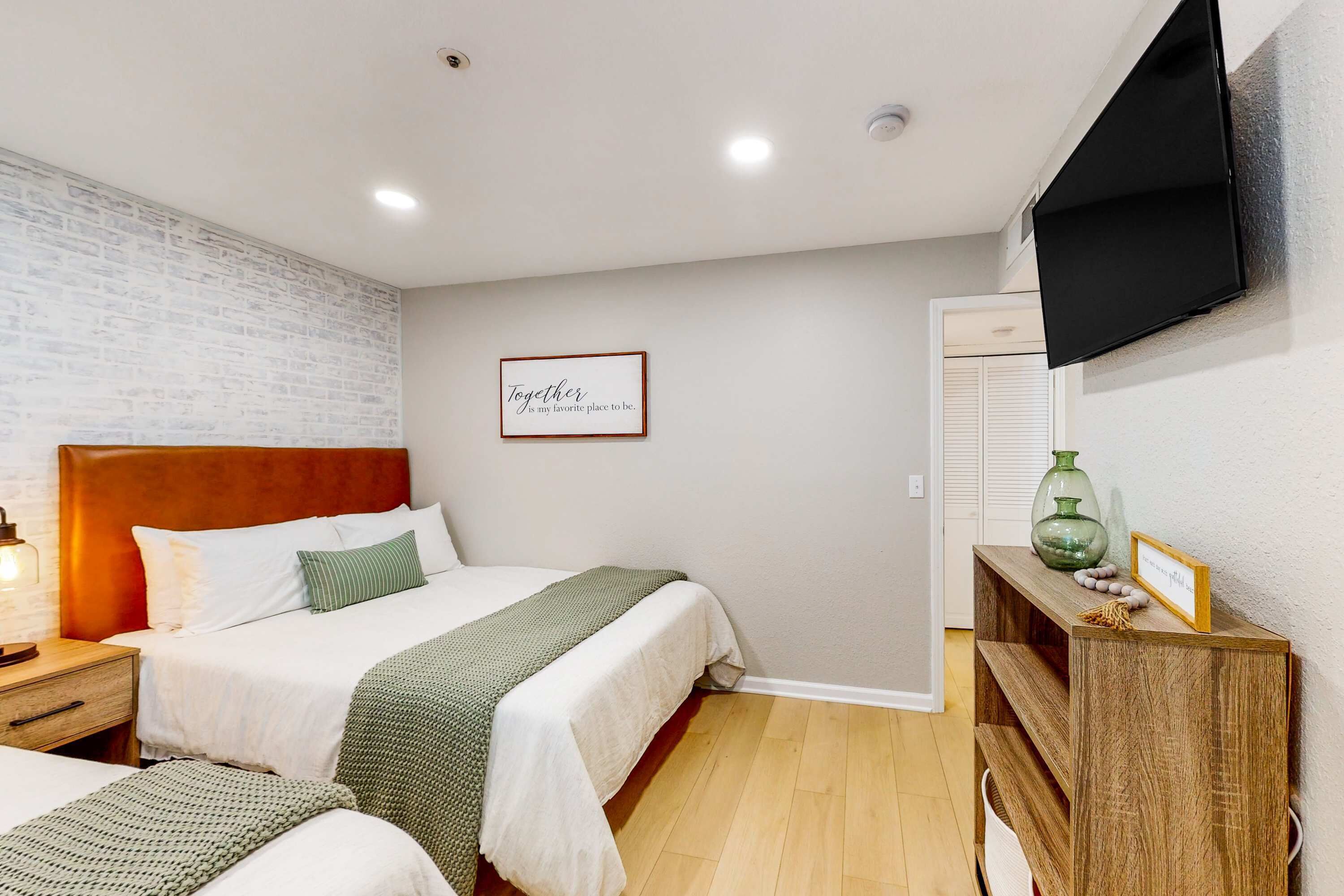 A hotel room with a brick-patterned wall, a wooden headboard, a bed with white bedding, a bedside table with a lamp, a wooden console table with decorative items, and a mounted TV.