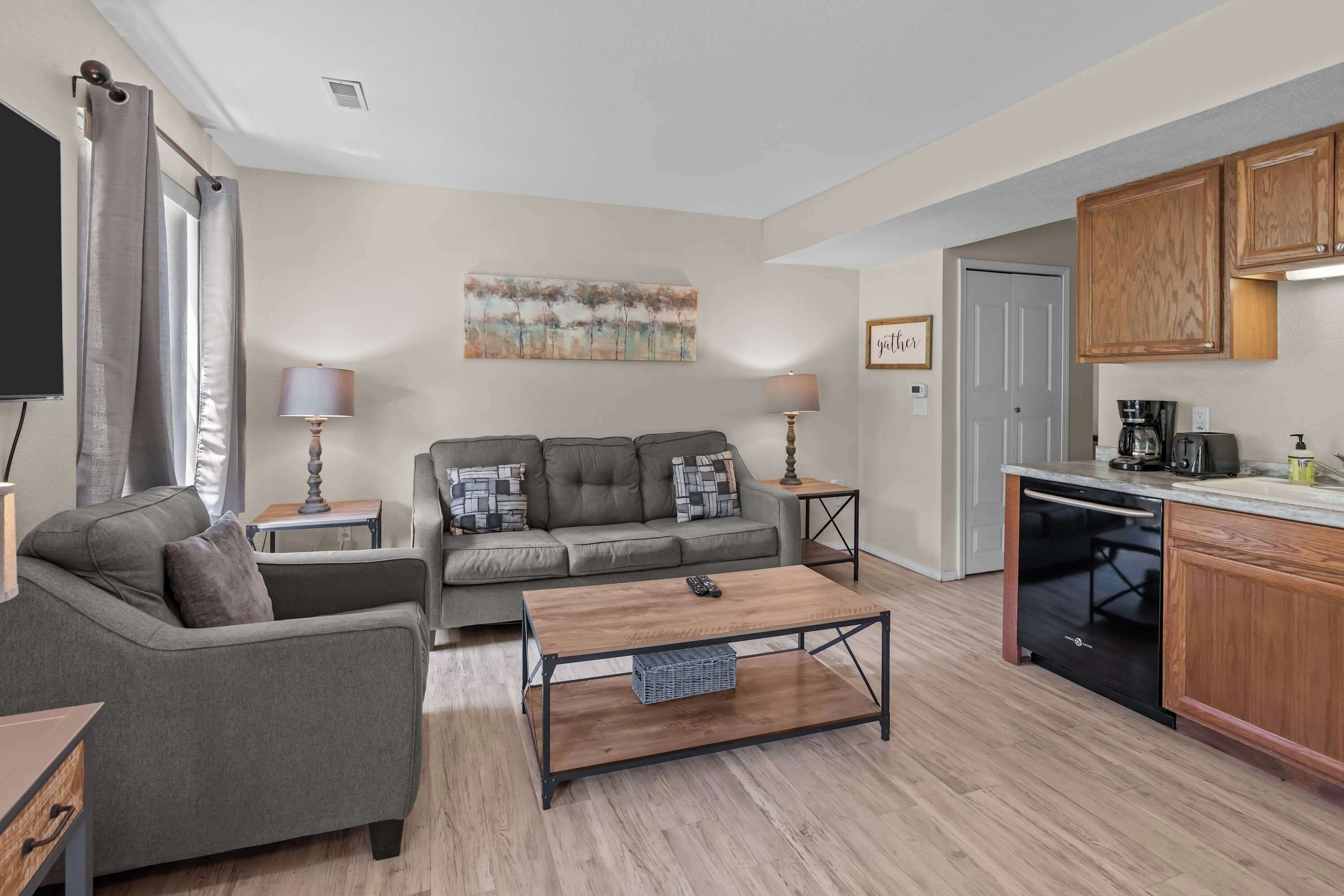 A modern living room with a grey sofa, a coffee table, and a kitchen area with wooden cabinets and a dishwasher.