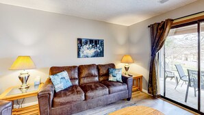 A living room with a leather sofa, two side tables with lamps, a painting on the wall, and a large sliding glass door leading to an outdoor area. - Rustic 2BR Condo - POOL & GOLF - Pet Friendly (Branson)