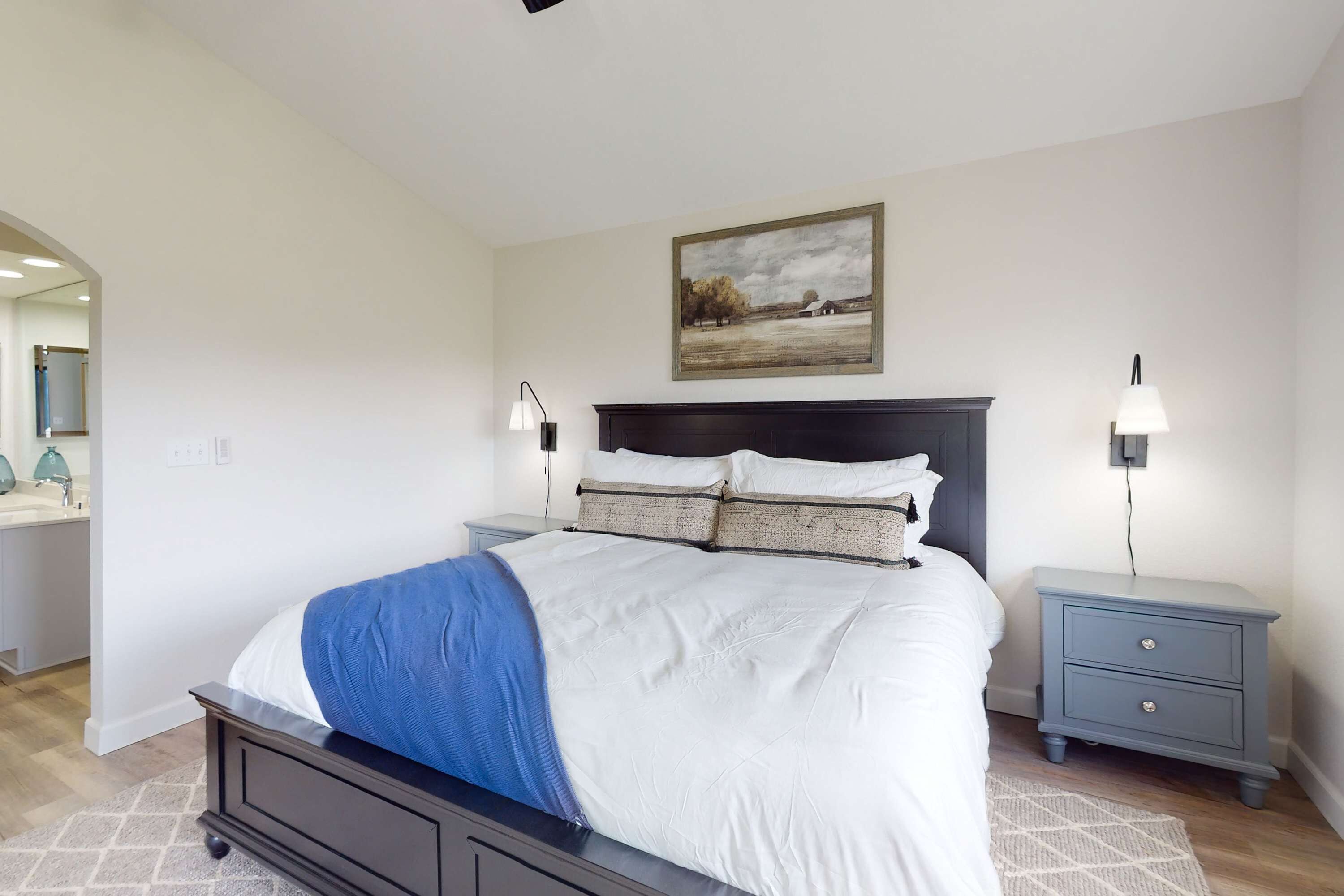 Une chambre à coucher avec un lit, des tables de chevet, une commode, un tableau et une salle de bain visible au fond.