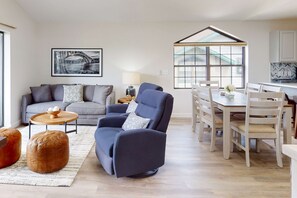 A modern living room with a sofa, armchair, ottoman, dining table, chairs, and a kitchen area with a window. - Waterview Oasis 2BR # Views # Pool & Fireplace (Branson)