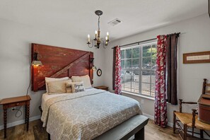 Un dormitorio con cabecera de madera, cama con ropa de cama estampada, un banco, una mesita de noche, una lámpara de pared y una ventana con cortinas.