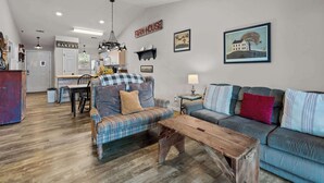 A spacious living room with a sofa, a coffee table, and a dining area with a kitchen in the background. - Rustic 2BR Condo - FARMHOUSE STYLE - Fall Creek (Branson)