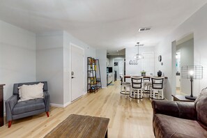 Ein Wohnzimmer mit grauem Sessel, braunem Ledersofa, Couchtisch aus Holz und einem Essbereich mit weißem Tisch und Stühlen.