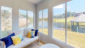 A room with a large window overlooking a body of water, a wicker sofa with cushions, and a view of a gazebo.