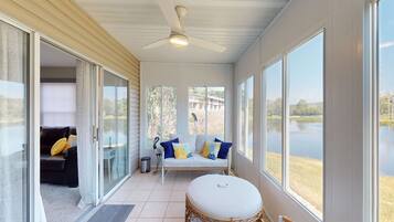 A room with a ceiling fan, a sofa with cushions, a round table, and large windows with a view of a lake.