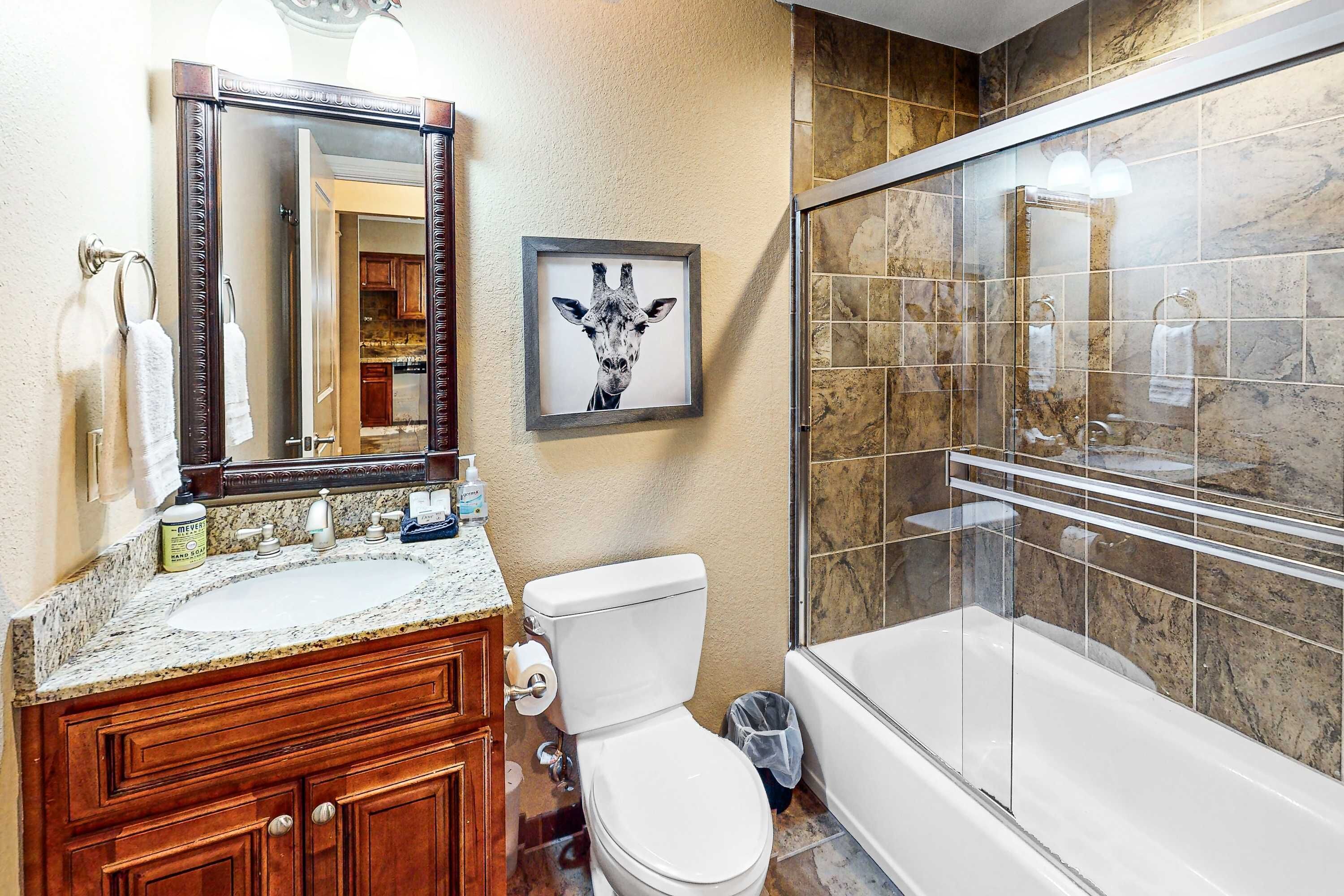 A bathroom with a marble shower, a framed giraffe picture, a wooden vanity, and a toilet.