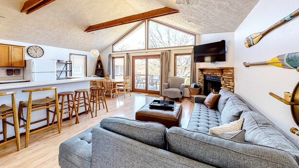 A spacious living area with a large sofa, wooden chairs, a dining table, and a kitchen area with a TV and wooden beams. - Lakefront 4BR Cabin # POOL TABLE # Lake Views (Reeds Spring)