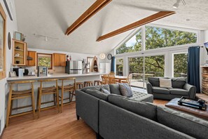 Ein geräumiger Wohnbereich mit einem großen grauen Sofa, Holzstühlen und einem Esstisch. Eine Küche mit Holzausbau und einer weißen Arbeitsplatte ist sichtbar. Große Fenster sorgen für viel Tageslicht und einen Ausblick ins Freie.