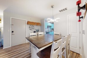 A dining area with a wooden table and white chairs, a kitchen in the background, and a ceiling light fixture. - Bright 3BR Condo - MOUNTAIN VIEWS - Community Pool (Branson)