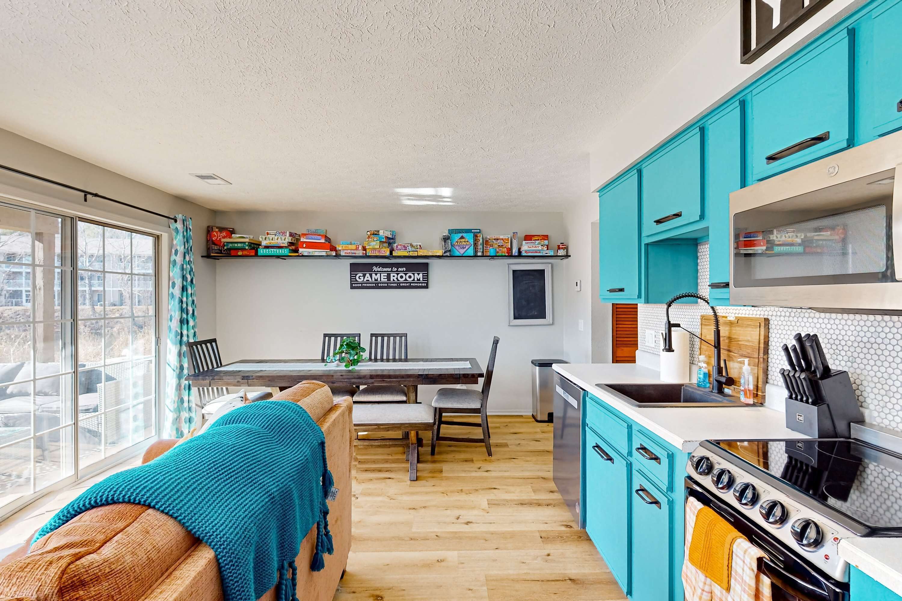 Une cuisine moderne avec des meubles de couleur turquoise, une table à manger en bois et une vue dégagée sur l’extérieur grâce à une grande fenêtre.