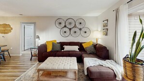 A living room with a brown sofa, a white ottoman, and a wall-mounted bicycle wheel decoration. - Serene 3BR Condo - MOUNTAIN VIEW - Community Pool (Branson)