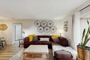 A living room with a brown sofa, a white ottoman, and a wall-mounted bicycle wheel decoration. - Serene 3BR Condo - MOUNTAIN VIEW - Community Pool (Branson)