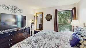 A bedroom with a flat-screen TV, a dresser, a bed with a floral bedspread, a mirror, a clock, and a window with curtains. - Serene 1BR Retreat - GOLF VIEW - Pools & Fireplace (Branson)