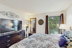 A bedroom with a flat-screen TV, a dresser, a bed with a floral bedspread, a mirror, a clock, and a window with curtains. - Serene 1BR Retreat - GOLF VIEW - Pools & Fireplace (Branson)