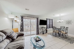 A living room with a sofa, a glass coffee table, a dining table with chairs, a TV, and a patterned curtain. - Inviting 1BR Condo - PET-FRIENDLY - Ground Floor (Branson)