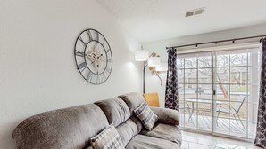 Een woonkamer met een bank, een wandklok, een vloerlamp en uitzicht door een schuifdeur.