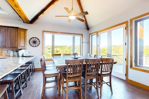 A spacious dining area with a large wooden table and chairs, a ceiling fan, and large windows offering a view of the outdoors. - Rustic 5BR Sanctuary - HOT TUB - Lake Access (Branson)