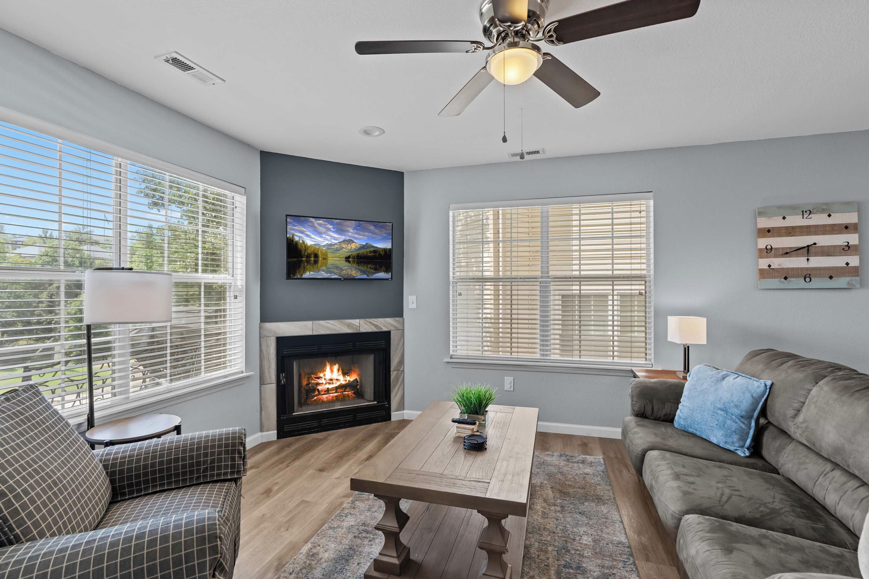 A living room with a fireplace, a television mounted on the wall, a ceiling fan, and large windows with blinds.