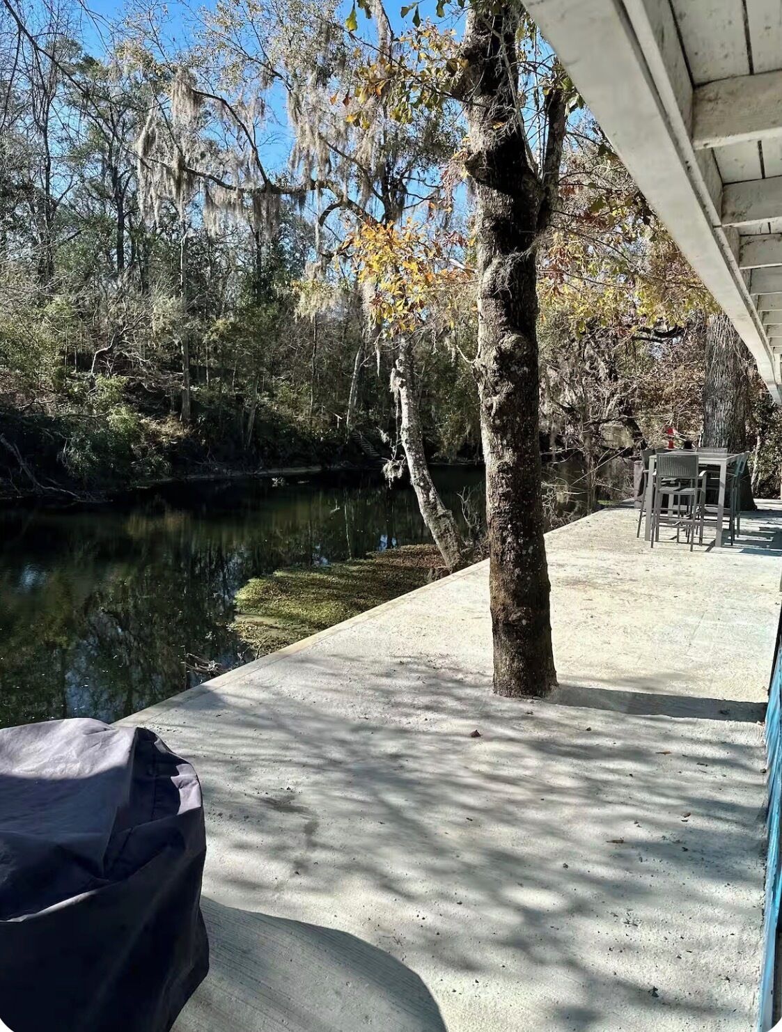 Una terraza de concreto con una mesa y sillas, con vista a un río tranquilo y árboles con hojas otoñales.