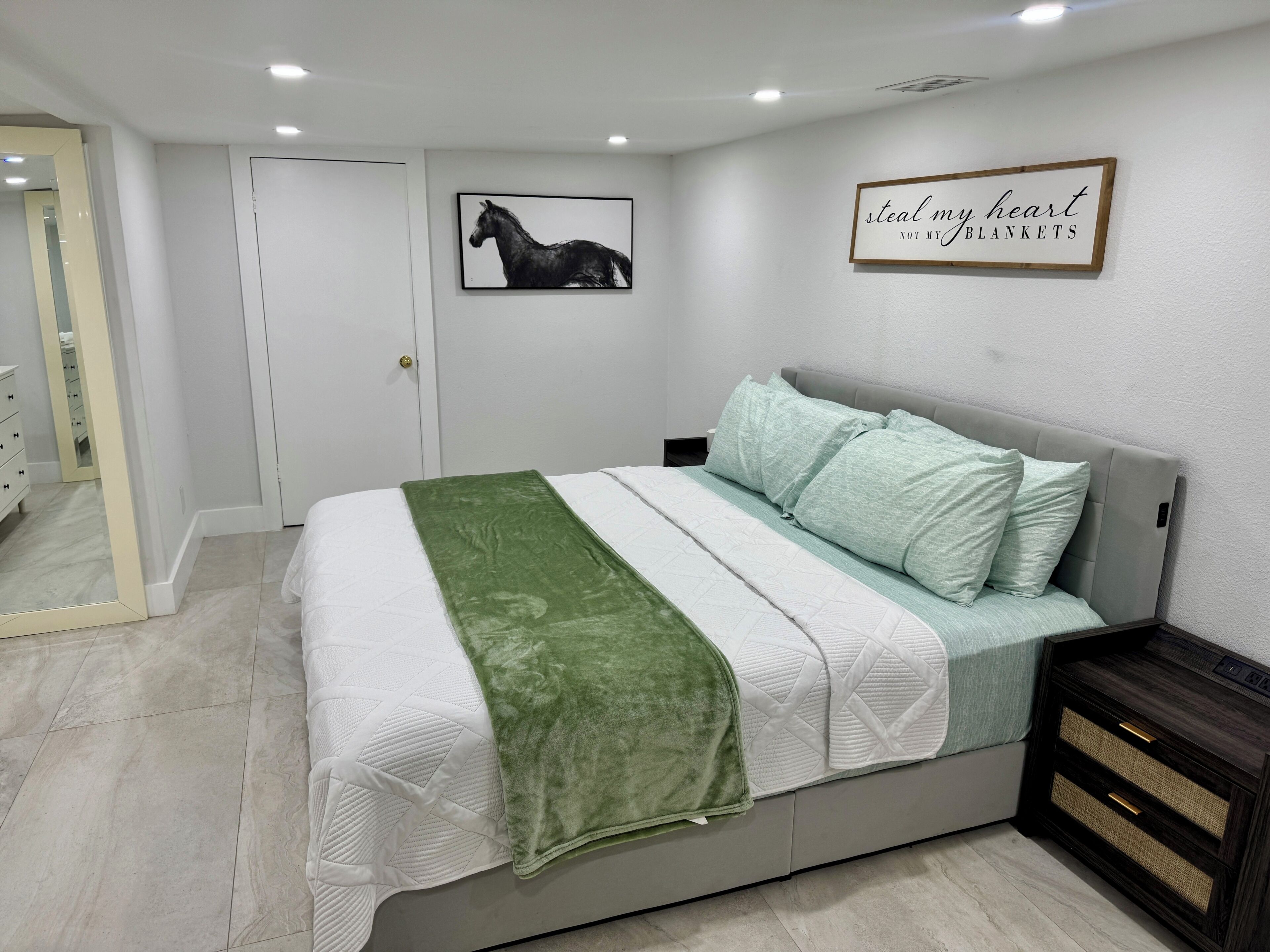 Ein Schlafzimmer mit einem Bett, einer grünen Tagesdecke, einem Kopfteil, einem Nachttisch, einem Wandbild eines Pferdes, einer Tür und einem Schild mit der Aufschrift 'Steal my heart not my blankets'.
