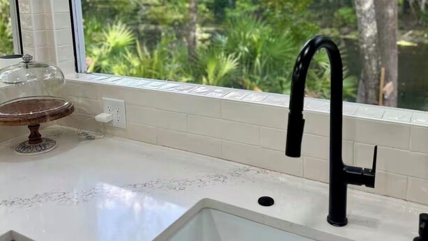 Una cocina con encimera blanca, grifo negro y ventana con vistas a un bosque.