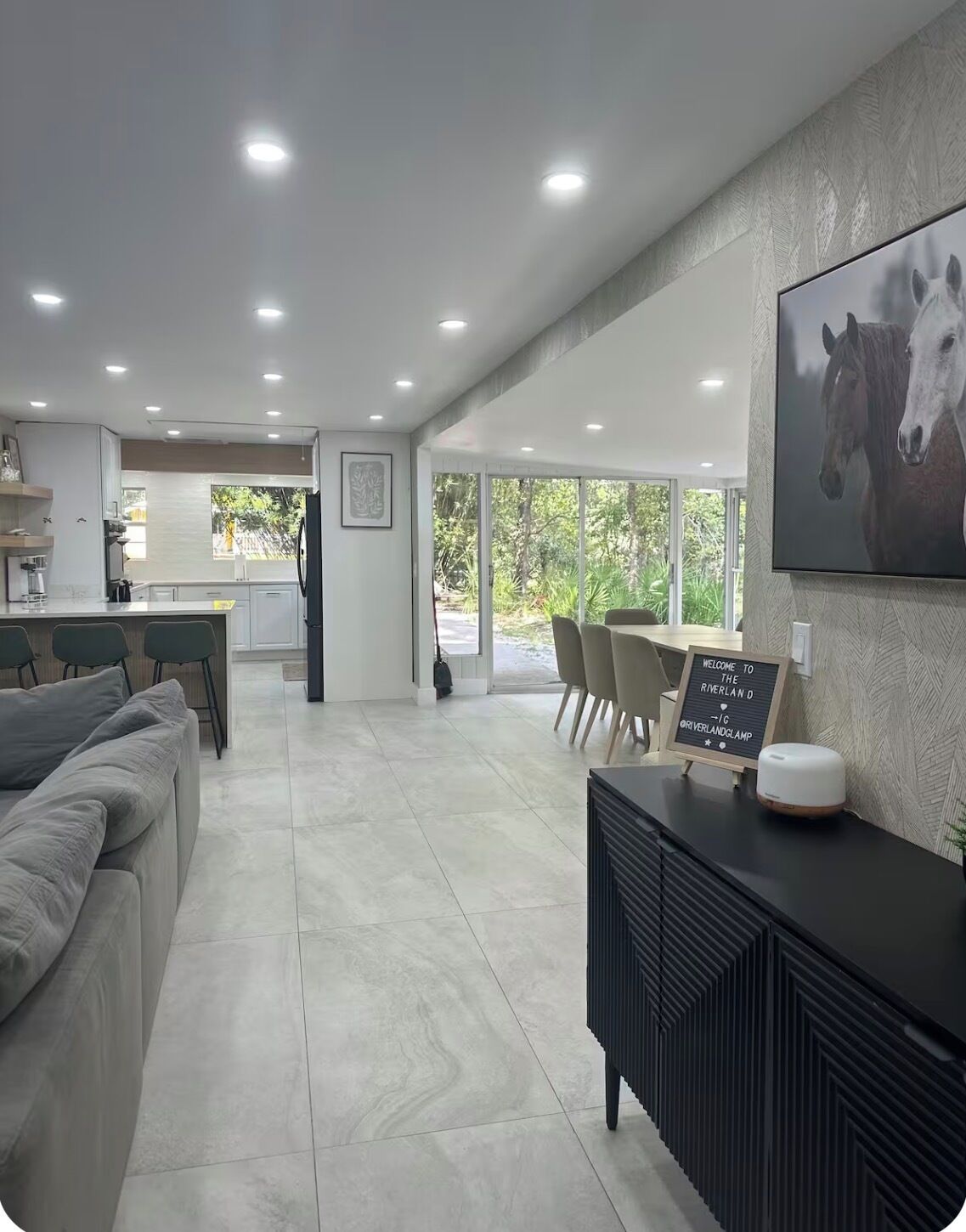 A modern kitchen with recessed lighting, a dining area with a table and chairs, and a large window offering a view of greenery.