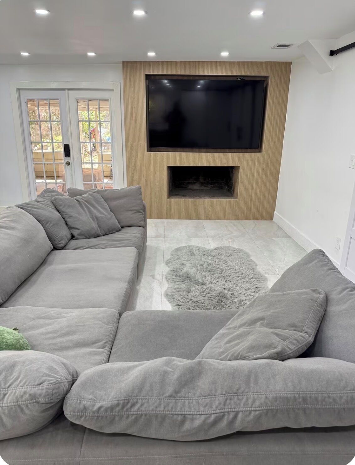 A modern living room with a sectional sofa, a flat-screen TV mounted above a fireplace, and a large window with a view of the outdoors.