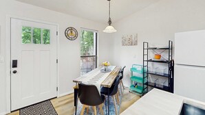 A compact kitchen with a dining table, chairs, a hanging light fixture, a door with a window, and a shelving unit with storage containers. - Rustic 2BR Cabin *POOL* Wooded (Kimberling City)