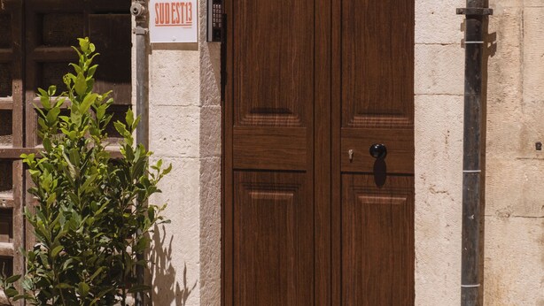A double wooden door with a sign above it, a number plaque, and a planter with red flowers. - Southeast 13 - Moon House (Putignano)
