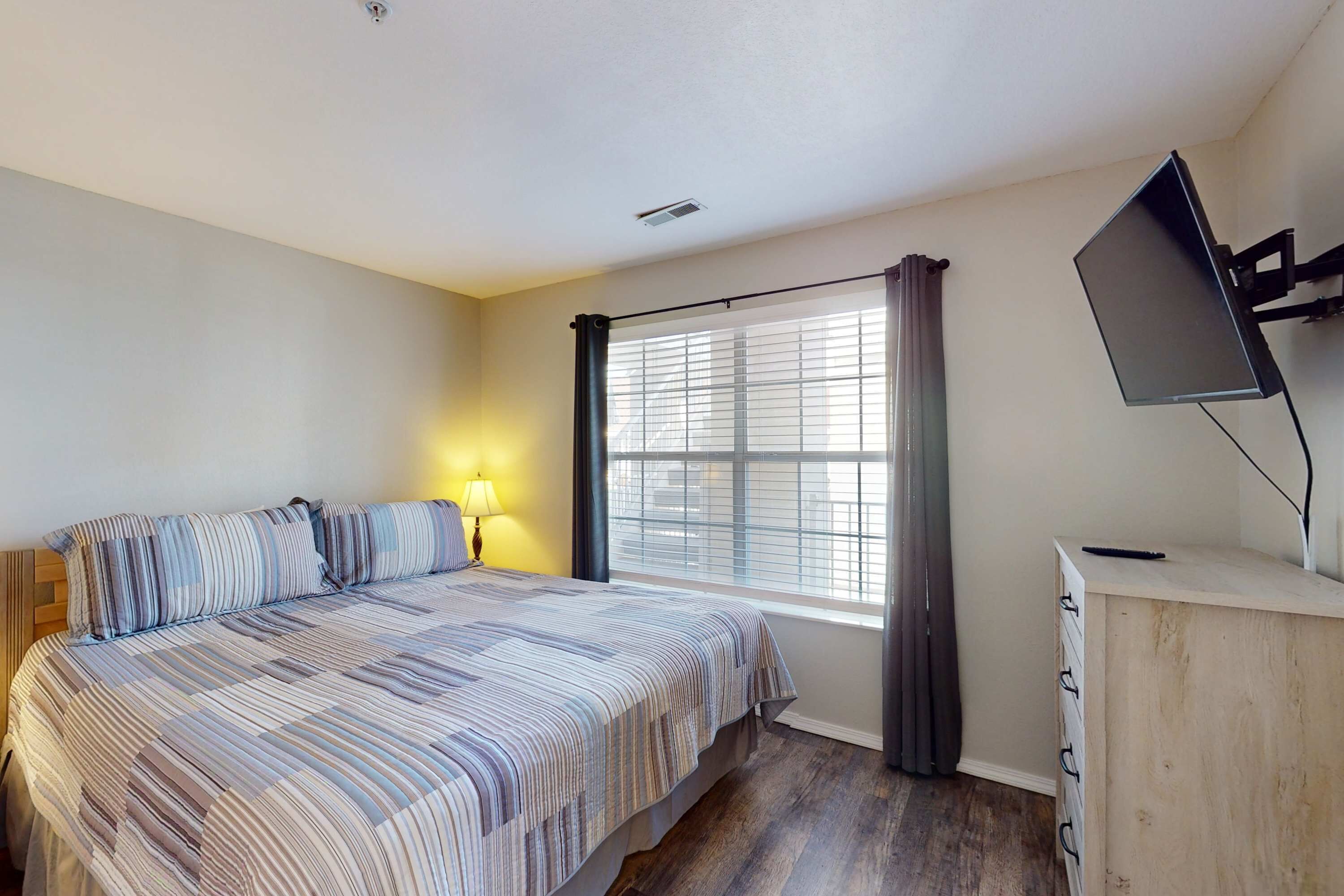 A bedroom with a bed, a desk, a TV, and a window with curtains.