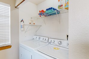 Ein Waschbereich mit zwei übereinander gestapelten Waschmaschinen und Trocknern, einem Regal mit Reinigungsmitteln und einem Fenster mit Jalousien.