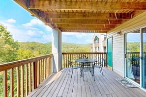 Eine Holzdiele mit Tisch und Stühlen, eine Glastür, die auf einen Balkon führt, und Blick auf einen bewaldeten Hügel.