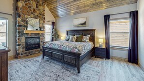 Ein Schlafzimmer mit Kamin aus Stein, einem Bett mit floralem Bettzeug, einer Holzbalkendecke und einem Fenster mit Vorhängen.